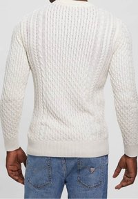 Crème-kleurige kabelgebreide trui met een textuurdesign, geribde boorden en onderrand, gedragen met blauwe spijkerbroek, toont een gelaagd breipatroon.
