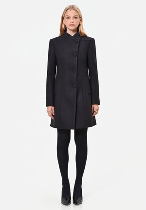 Donna in piedi che indossa un cappotto nero aderente al ginocchio con grandi bottoni, collant neri, tacchi neri e orecchini dorati su sfondo bianco.