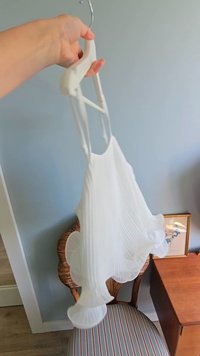 Bílá plisovaná šaty zavěšená na ramínku, s tenkými ramínky a rozšířeným lemem. Pozadí zahrnuje dřevěnou židli a zarámované umělecké dílo.