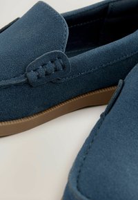 Mocassins en suede bleu avec une texture lisse, semelle en caoutchouc marron contrastante et couture décorative sur le dessus pour un détail supplémentaire.