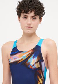 Costum de baie în albastru și negru cu modele abstracte; prezintă un decolteu înalt, curele subțiri și detalii cu logo pe piept.