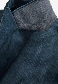 Marineblaue Jacke mit einem strukturierten geometrischen Muster, einem dunkelgrauen Filzkragen und kontrastierenden Nähten entlang der Kanten. Mit Reißverschluss vorne.