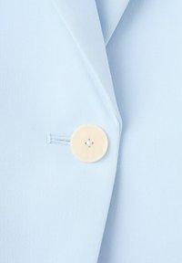 Lichtblauw stof met een enkele cremekleurige ronde knoop, genaaid vlakbij een verticale knoopsgat aan de rand van een kledingstuk.