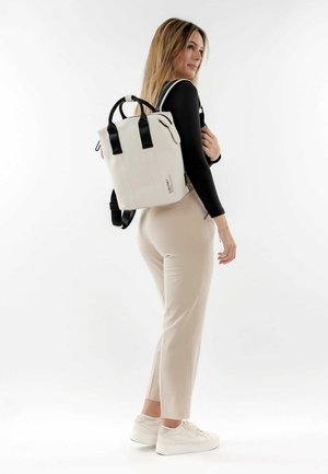 Weißer Rucksack mit schwarzen Trägern, rechteckige Form, glatte Textur, mit Logodetail an der Seite, von einem Model auf den Schultern getragen.