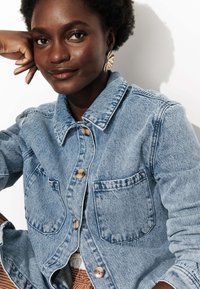 Femme souriante avec des cheveux courts et bouclés porte des boucles d'oreilles en or en forme d'éventail et une veste en denim bleu clair avec de grandes poches avant et des boutons marron.