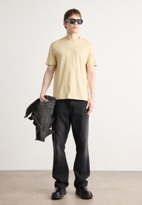 Lys gul t-shirt, sorte jeans, sorte solbriller og en denimjakke. Modellen står med jakken over den ene arm, iført kraftige sorte sko.