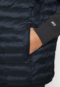 Ręka wsuwająca się do zamkniętej kieszeni granatowej pikowanej kurtki noszonej na ciemnej bluzie z długim rękawem z małym logo flagi na rękawie.