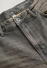 Gros plan d’un jean gris foncé avec un bouton, une passoire pour ceinture, des poches cousues et l’inscription « Relaxed Tapered Fit » imprimée à l’intérieur de la ceinture.
