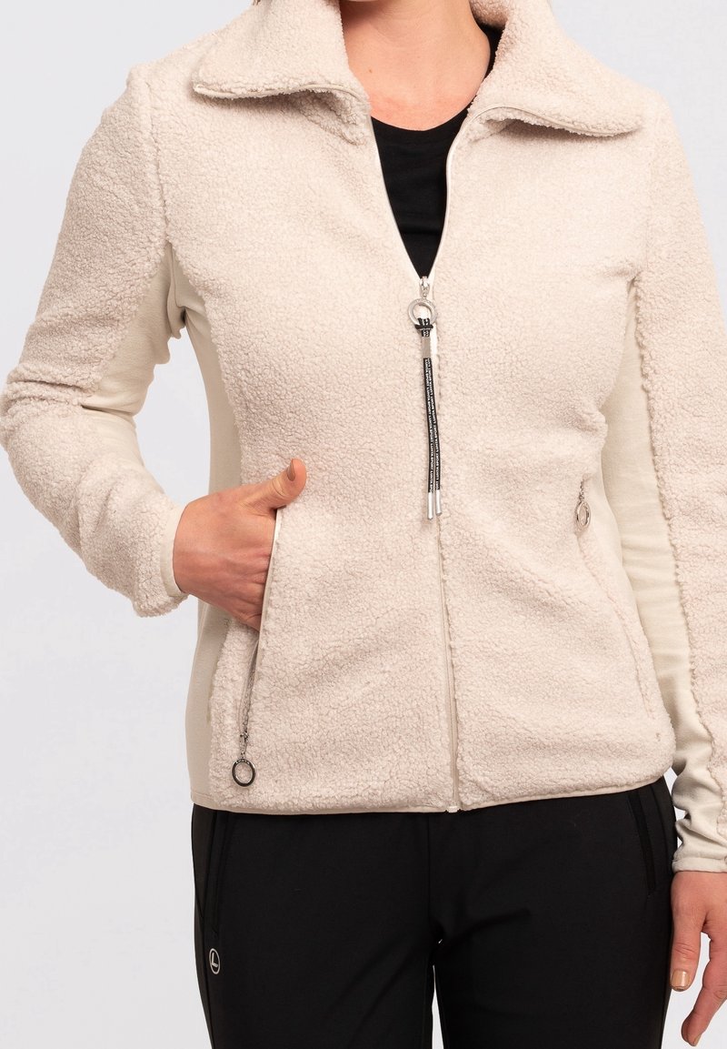 Femme portant une veste polaire beige zippée avec des poches latérales et un pantalon noir, une main dans la poche, sur un fond clair uni.