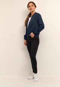 Veste de pilote marine avec fermeture éclair à l'avant, poignets et ourlet côtelés, associée à un pantalon skinny noir et des baskets blanches. Le modèle se tient devant un arrière-plan neutre.