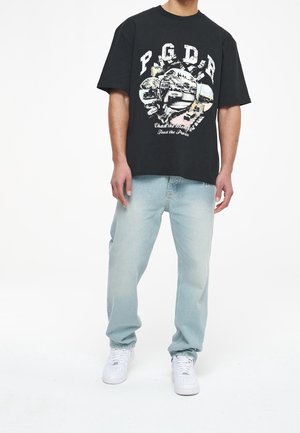 Homme portant un t-shirt noir à motif avec texte et un jean bleu clair, debout sur un fond blanc avec des baskets blanches.