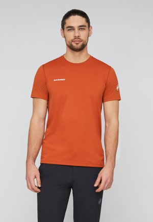 Mand med mørkt hår og skæg iført en orange Mammut t-shirt og sorte bukser, der står mod en ensfarvet grå baggrund.