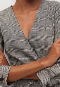Szara bluzka w kratę z zapięciem w kształcie litery V. Posiada krótkie rękawy, teksturowany materiał oraz czarne guziki na mankietach.