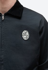 Μαύρο μπουφάν με φερμουάρ μπροστά, γιακά από κορντόρι και οβάλ ψηφιδωτό με έναν ιππότη πάνω σε άλογο που κρατά δόρυ και τη λέξη "ΠΑΛΑΙΟΣ".