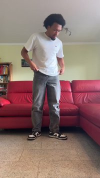 Λευκό κοντομάνικο μπλουζάκι, γκρι τζιν παντελόνι και μαύρα αθλητικά παπούτσια με λευκές λεπτομέρειες. Στέκεται σε ένα δωμάτιο με κόκκινο καναπέ και ανοιχτόχρωμους τοίχους.