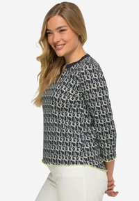 Blusa a fantasia navy con maniche a tre quarti, dotata di scollatura a V, bordo chiaro a contrasto e orlo ondulato. Il tessuto sembra morbido e leggero.