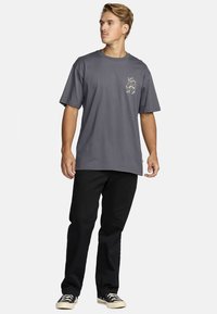 Hall halli puuvillane T-särk lühikeste varrukatega ja taskuga, millel on graafiline kujundus. Kanditud mustade pükstega ja mustade tossudega.