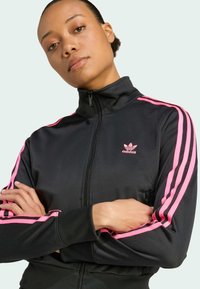 Donna con le braccia incrociate che indossa una giacca a track nera di Adidas con strisce rosa e il logo sulle maniche e sul petto, su uno sfondo semplice.