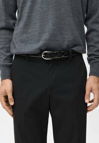 Uomo che indossa una maglia a maniche lunghe grigio scuro infilata in pantaloni neri con cintura di pelle nera e fibbia d'argento, mani rilassate ai lati.