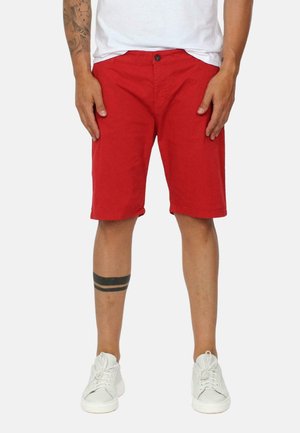 Pantaloni corti rossi in cotone con chiusura a bottone, taglio dritto e tasche laterali. Indossati con scarpe da ginnastica bianche e una maglietta bianca semplice.