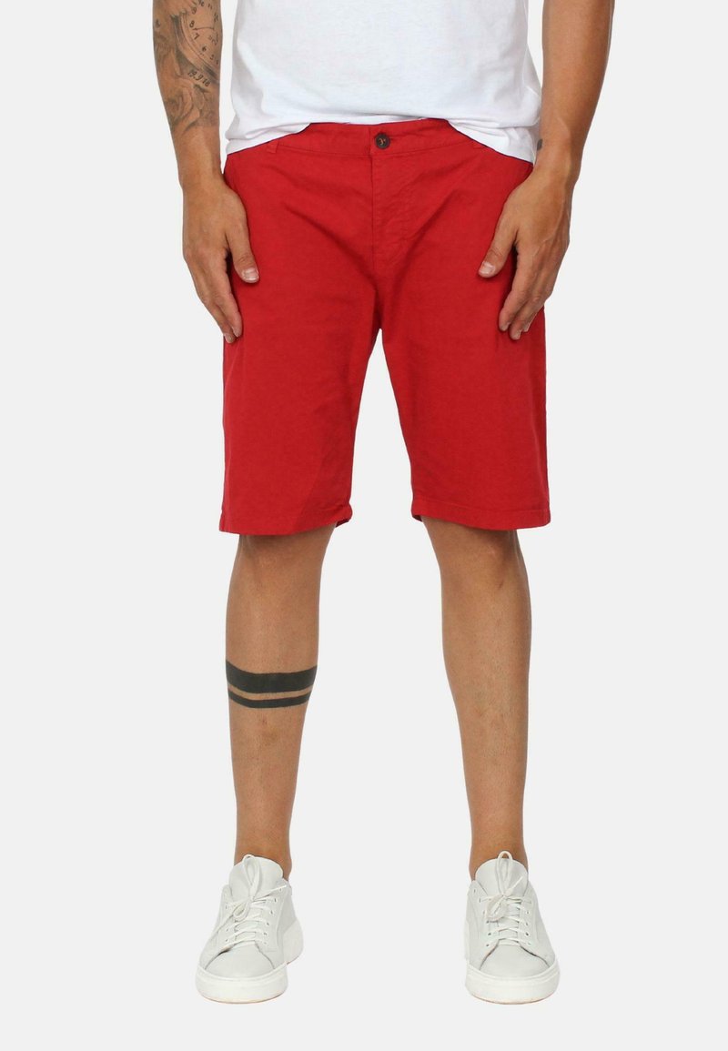 Pantaloni corti rossi in cotone con chiusura a bottone, taglio dritto e tasche laterali. Indossati con scarpe da ginnastica bianche e una maglietta bianca semplice.