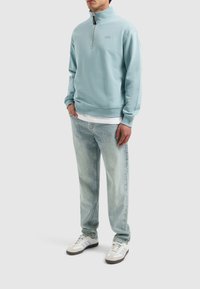 Lichtblauw sweatshirt met ritssluiting van zacht materiaal, gecombineerd met vervaagde denim jeans en witte sneakers met grijze accenten.