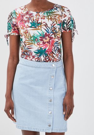 Vrouw met een bloemenprint korte mouw top met gebonden mouwen en een lichtblauwe spijkerrok met knopen aan de voorkant.