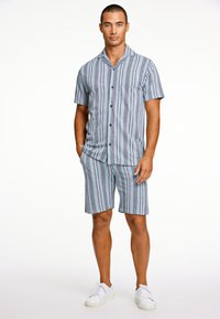 Camicia a maniche corte a righe e pantaloni corti coordinati in tonalità di blu, con chiusura a bottoni sul davanti e vestibilità rilassata, abbinati a sneaker bianche.