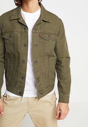 Muž v olivově zelené denimové bundě přes bílé tričko a béžové kalhoty, stojící na jednoduchém světle zbarveném pozadí.