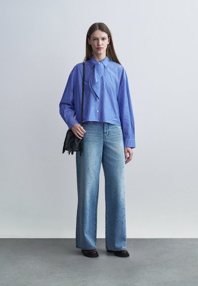 Jeune femme debout portant un chemisier à rayures bleues, un jean large orné de petites décorations, des mocassins noirs et un sac à bandoulière noir avec des franges.