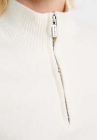 Cream-colored sweater with a ribbed high neck and a half zipper. Soft texture, lightweight, featuring a small branded zipper pull accent.