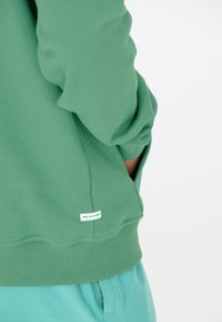 Grüner Pullover mit gerippten Bündchen, strukturiertem Stoff und einem kleinen Etikett mit der Aufschrift "NEW BAD HABITS" nahe dem Saum. Kombiniert mit türkisfarbenen Hosen.