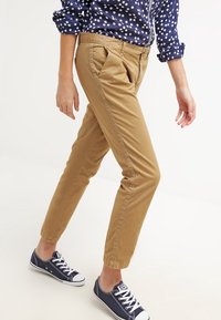 Chinos kaki coupe slim, avec des plis à l'avant, des poches latérales et un ourlet retroussé, associés à un chemisier bleu marine à pois et des baskets.