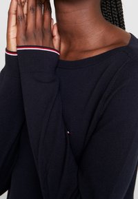 Pull en maille marine avec un col large et des accents rayés rouges et blancs sur les poignets. La texture est lisse et respirante.