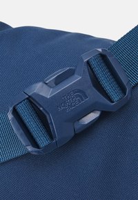 Fibbia in plastica blu con il logo "The North Face" che fissa una cinghia intrecciata blu su un tessuto texturizzato.