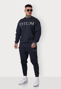 Sweat-shirt marine avec le logo "ESTEEM" en blanc, associé à un jogging assorti et des baskets blanches ; tissu lisse, coupe décontractée, poignets côtelés.