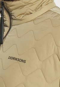 Jasny beżowy, pikowany płaszcz z wzorzystym pikowaniem. Posiada zamek błyskawiczny i czarne logo "DIDRIKSONS" z przodu.