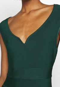 Bodysuit vert à côtes avec un décolleté en V profond. Le tissu a une texture lisse et une coupe structurée, mettant en valeur la taille.