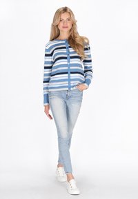 Blau gestreifter Cardigan mit Knopfleiste, hellblauer Hintergrund, mit dunkelblauen, marineblauen und weißen Streifen. Kombiniert mit hellblauen Jeans und Sneakers.