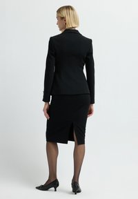 Blazer nero su misura con un design doppiopetto, abbinato a una gonna a matita fino al ginocchio con spacco sul retro. Calze e scarpe con tacco basso.
