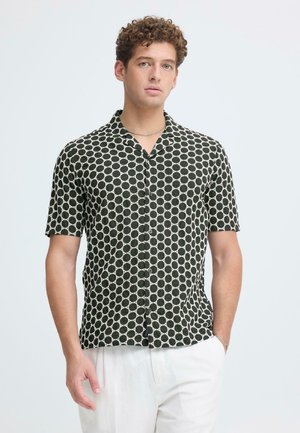 Mand med krøllet hår iført en sort kortærmet skjorte med hvide cirkelmønstre og hvide bukser, stående med den ene hånd i lommen.