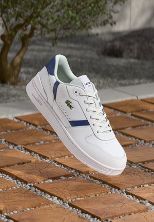 Biały sneaker Lacoste z niebieskimi akcentami i zielonym logo krokodyla, prezentowany na drewnianej podłodze z fugami wypełnionymi żwirem.