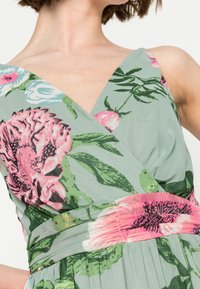 Žena v bezramínkových zelených šatech s růžovými květinovými vzory a zavinovacím designem, vyfotografována od krku po pas.