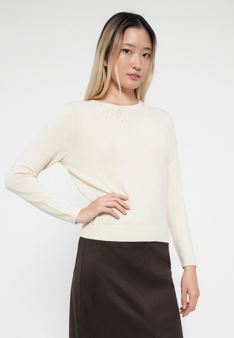 Suéter crema de manga larga con cuello redondo, que presenta sutiles adornos en la parte superior y puños acanalados, combinado con una falda oscura.