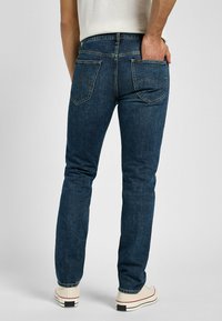 Jeans de mezclilla azul oscuro con corte slim, que cuentan con dos bolsillos traseros y detalles de costura. Los puños están ligeramente doblados y se combinan con zapatillas blancas.