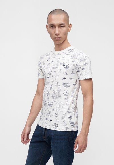 Άνδρας που φοράει λευκό εφαρμοστό T-shirt με ναυτικό τύπωμα σε ναυτικό μπλε και μπλε τζιν, στέκεται μπροστά σε απλό φωτεινό φόντο.