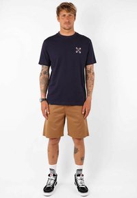 T-shirt bleu marine avec un petit design multicolore sur la poitrine, associée à un short beige, des baskets noires et des chaussettes blanches à motifs.