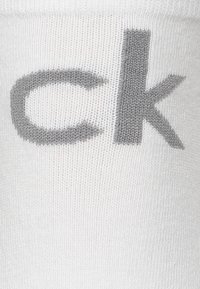 Balta kojinė su pilku "ck" logotipu. Audinys lygus ir megztas, su gurguliuotu raštu visame paviršiuje, užtikrinančiu tamprumą ir patogumą.