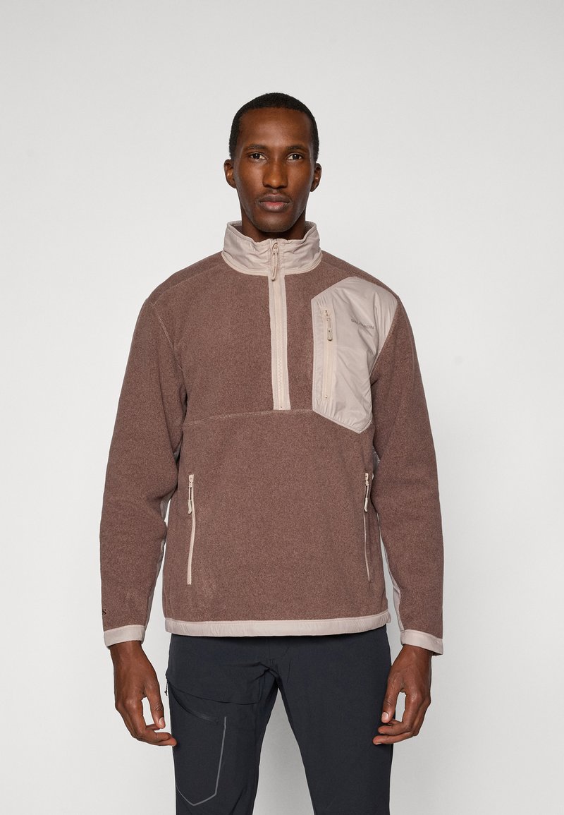 Pull en polaire marron avec un col montant, fermeture éclair, accents beige clair et poche poitrine. Comprend deux poches latérales zippées et des poignets élastiques.