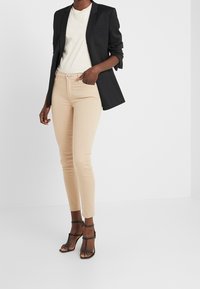 Blazer negro con solapas estructuradas, camiseta crema y jeans ajustados de color beige claro; combinado con sandalias de tacón negro con tiras.